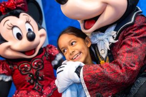 Child smiles as she hugs Mickey Mouse, with Minnie Mouse in the background.