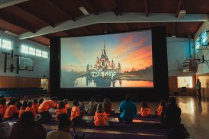 Kids in orange shirts watch a Disney movie on a large screen in a gym.