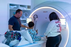 child entering MRI machine gives a thumbs up to his mom and technician