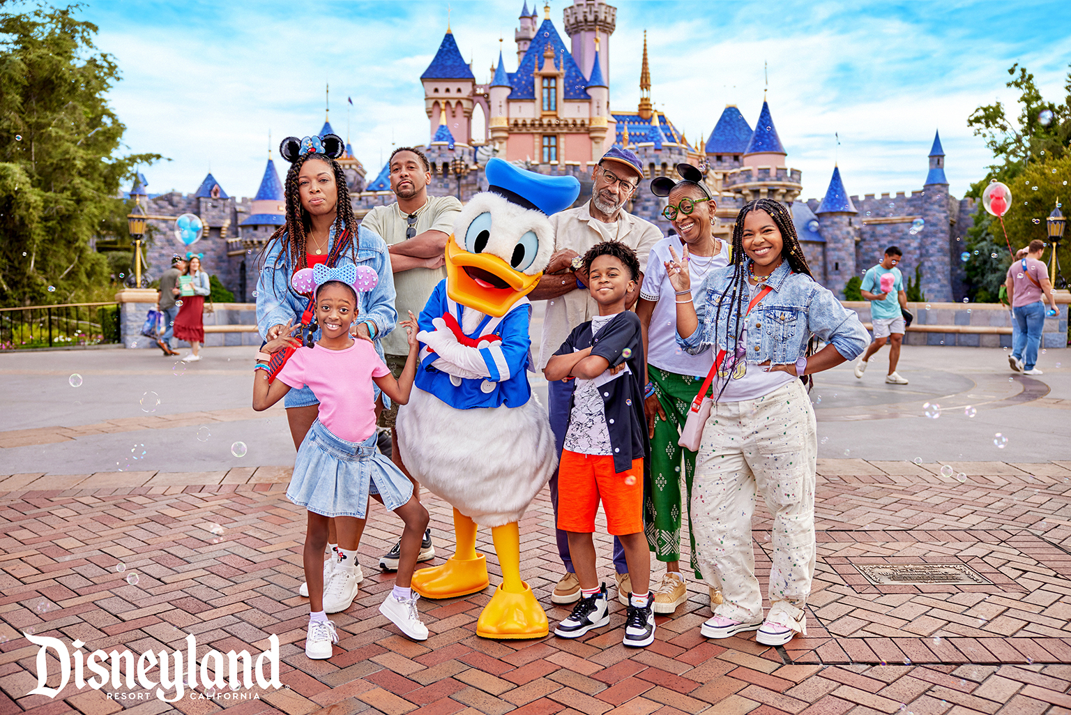 A Black family and Donald Duck all with their arms crossed posing for a picture in front of the Disneyland Resort castle.